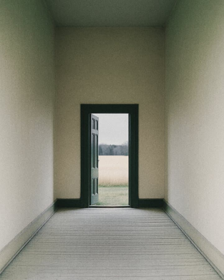 A bare corridor leading to an open door, with a field beyond